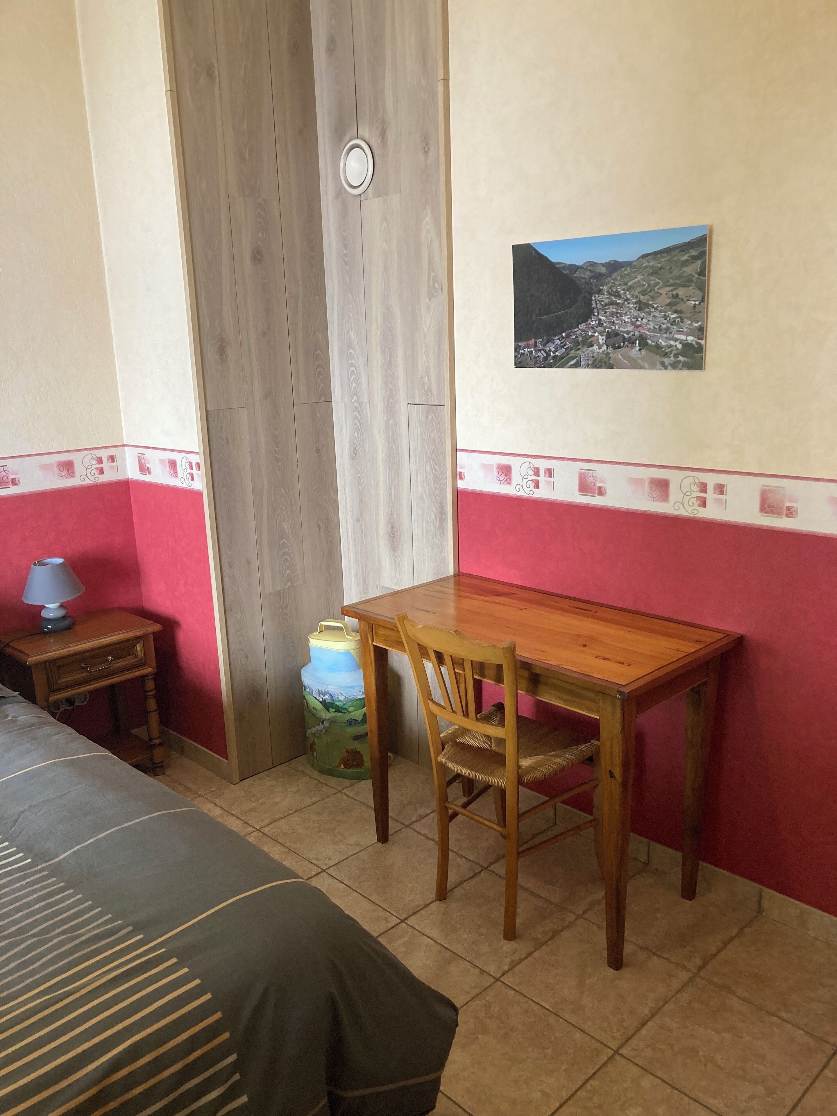 A room with a wooden table and chair, a bed with a striped cover, a small bedside table with a lamp, a large water dispenser, and a wall-mounted picture of a mountainous landscape.
