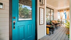 Une porte turquoise avec une fenêtre, une pancarte de bienvenue et du mobilier d’extérieur sur une véranda.