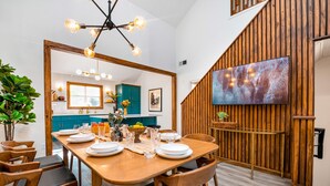 Un coin repas avec une table en bois dressée pour quatre personnes, un lustre moderne et un mur en bois sur lequel est fixée une télévision.
