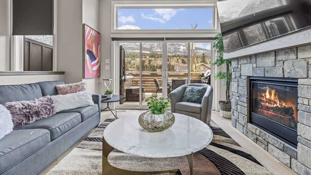A modern living room with a fireplace, a sofa, a coffee table, and a view of a mountainous landscape through large windows. - BRAND NEW|Hofburg by SSR|TopView|Luxury|PRIVATE HOT TUB|4BR plus Den ( BL# RES-11683 ) (Canmore)
