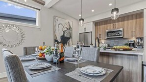 A modern dining area with a dark wooden table set for four, a large abstract painting on the wall, and a view of the kitchen in the background. - BRAND NEW|Hofburg by SSR|TopView|Luxury|PRIVATE HOT TUB|4BR plus Den ( BL# RES-11683 ) (Canmore)