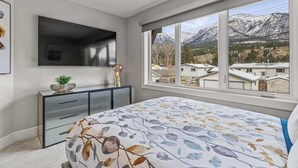 A bedroom with a bed, a TV mounted on the wall, a console table, and a large window with a mountain view. - BRAND NEW|Hofburg by SSR|TopView|Luxury|PRIVATE HOT TUB|4BR plus Den ( BL# RES-11683 ) (Canmore)