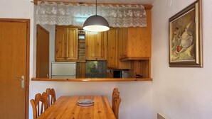 A wooden dining table with chairs, a kitchen with wooden cabinets, a pendant light, and a painting on the wall.