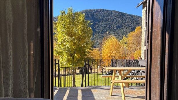 A room with a large window, a bed, and a view of a patio with a wooden deck, a metal railing, and autumn-colored trees.