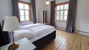 A bedroom with a bed, a bedside table with a lamp, two windows with curtains, and a hanging light fixture.