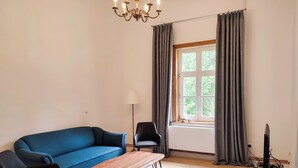 A living room with a blue sofa, a wooden coffee table, a black armchair, a floor lamp, a television on a stand, and a window with curtains.