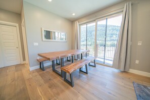 Ein geräumiger Raum mit einem großen Holztisch und Bänken, einer Glasschiebetür, die auf einen Balkon mit Bergblick führt, und einem gerahmten Spiegel an der Wand.