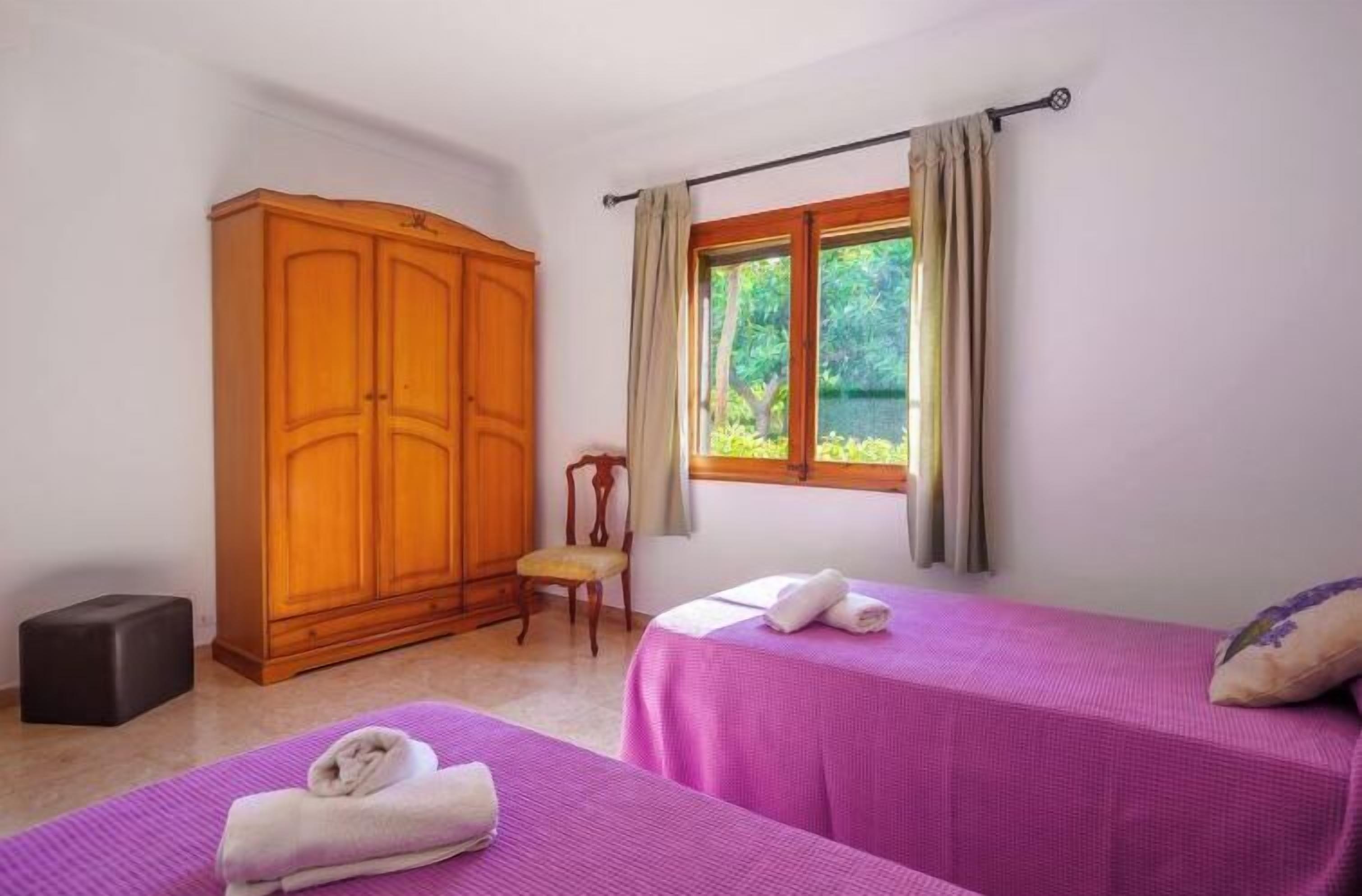 A room with two beds, a wooden wardrobe, a chair, and a window with curtains.