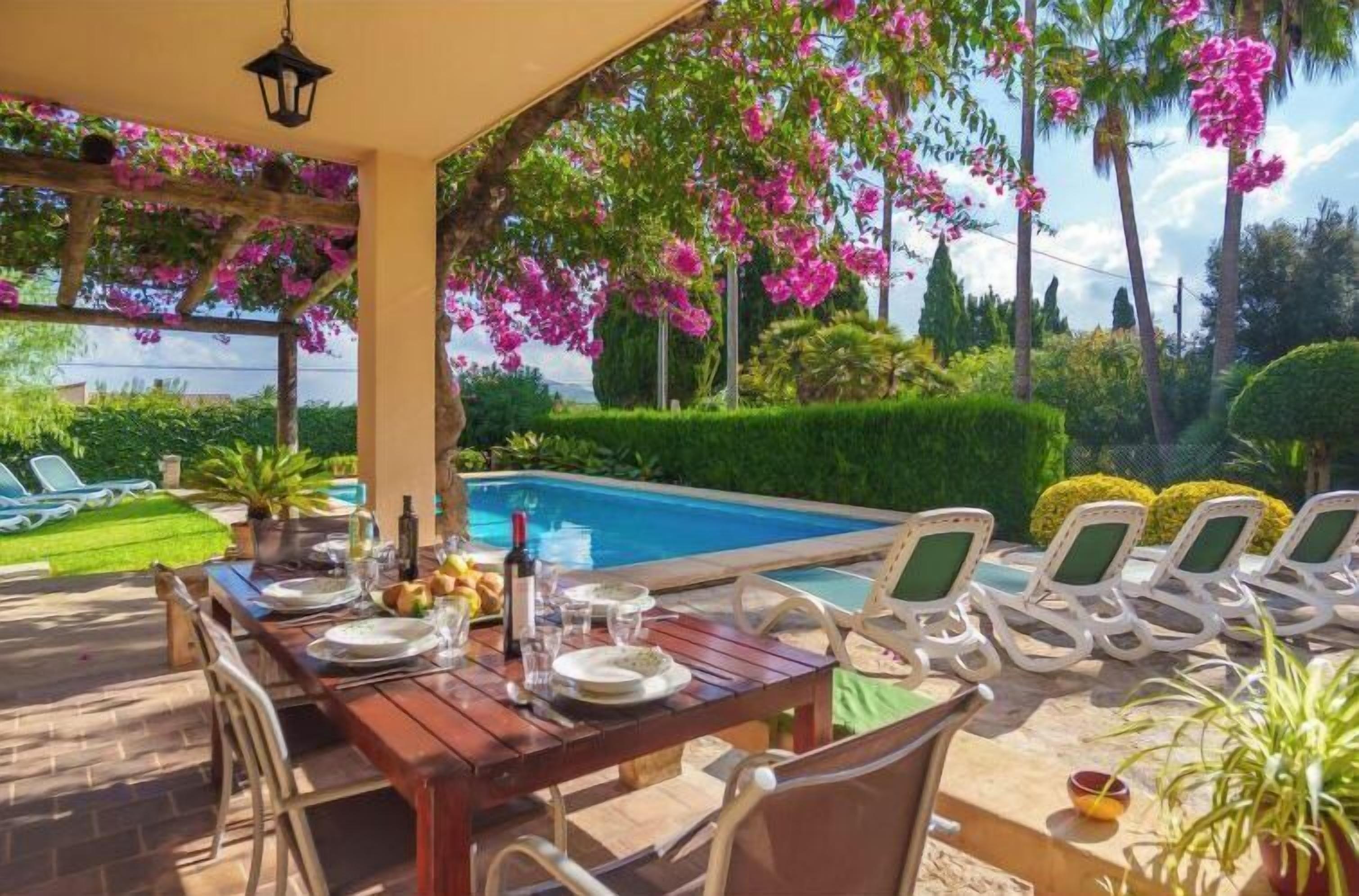 Un área de comedor al aire libre con vista a la piscina, mesa de madera, sillas y una pérgola con plantas florecientes.