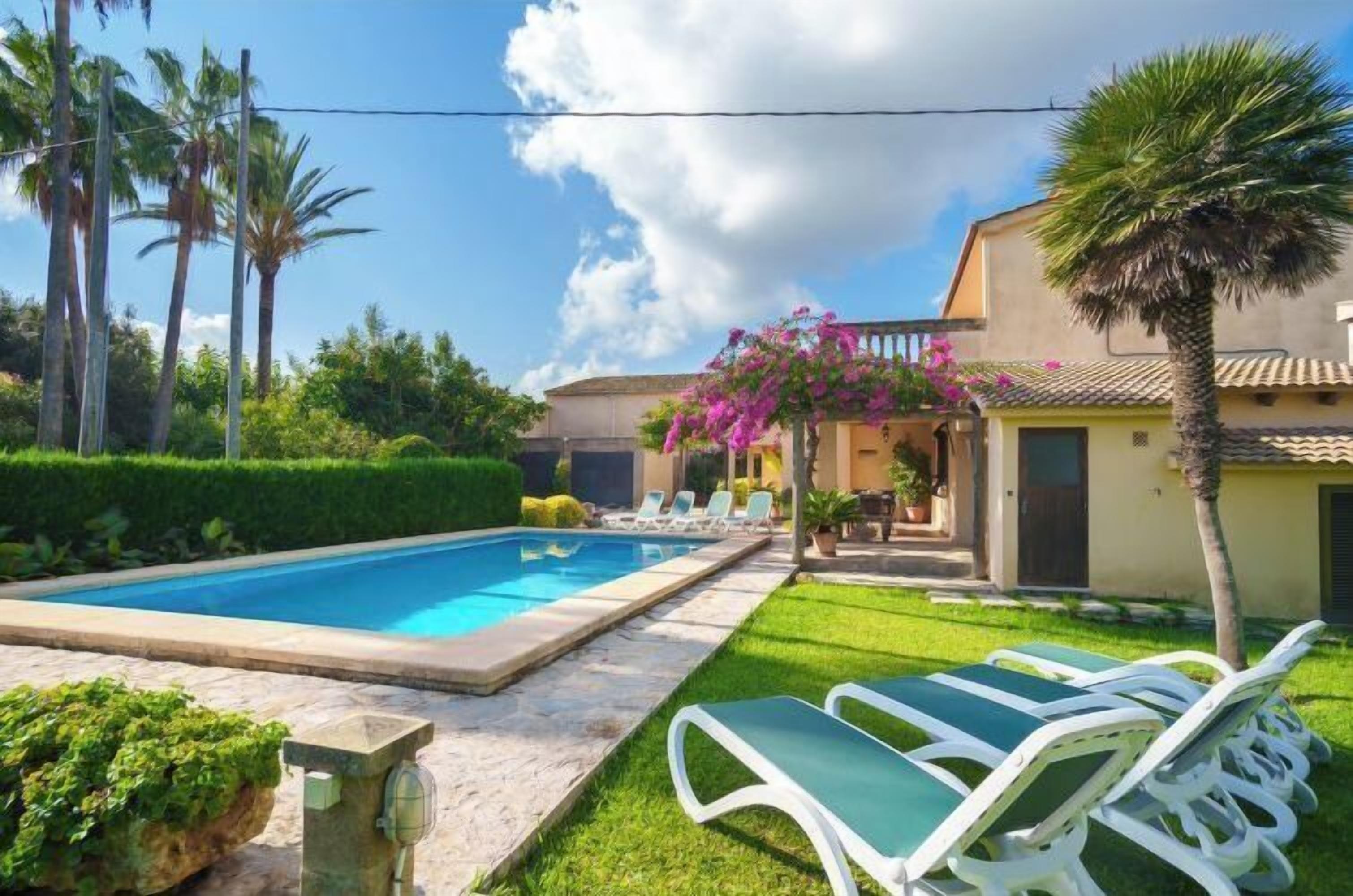 A swimming pool with lounge chairs, a garden with palm trees, and a building with a tiled roof.