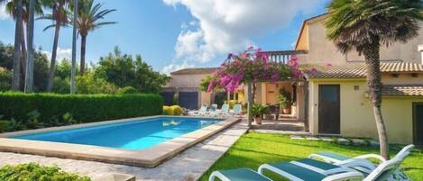 A swimming pool with lounge chairs, a garden with palm trees, and a building with a tiled roof.