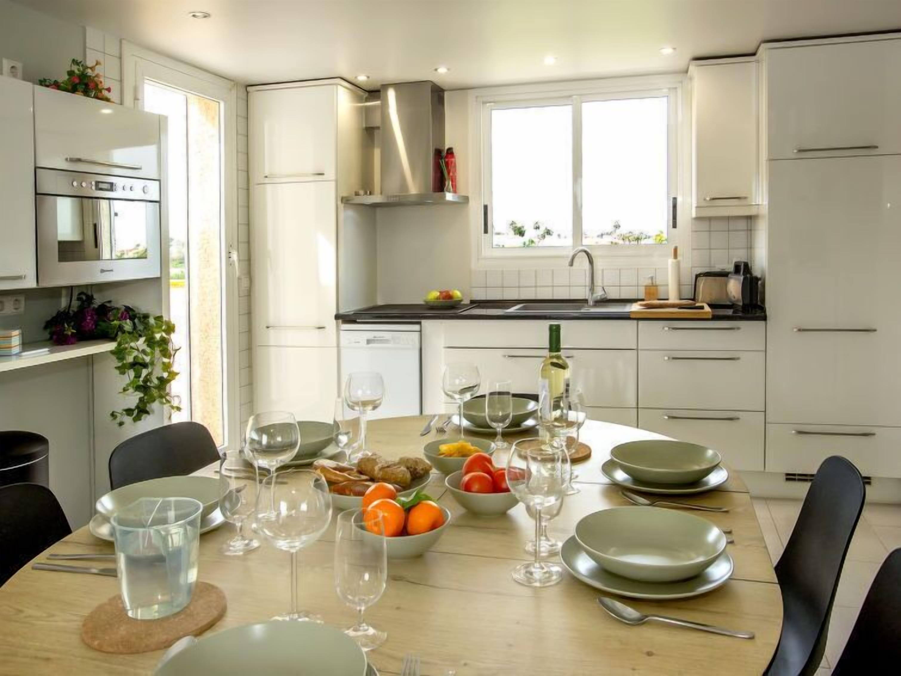 Une cuisine moderne avec des meubles blancs, une table à manger dressée avec des assiettes, des verres et une bouteille de vin.