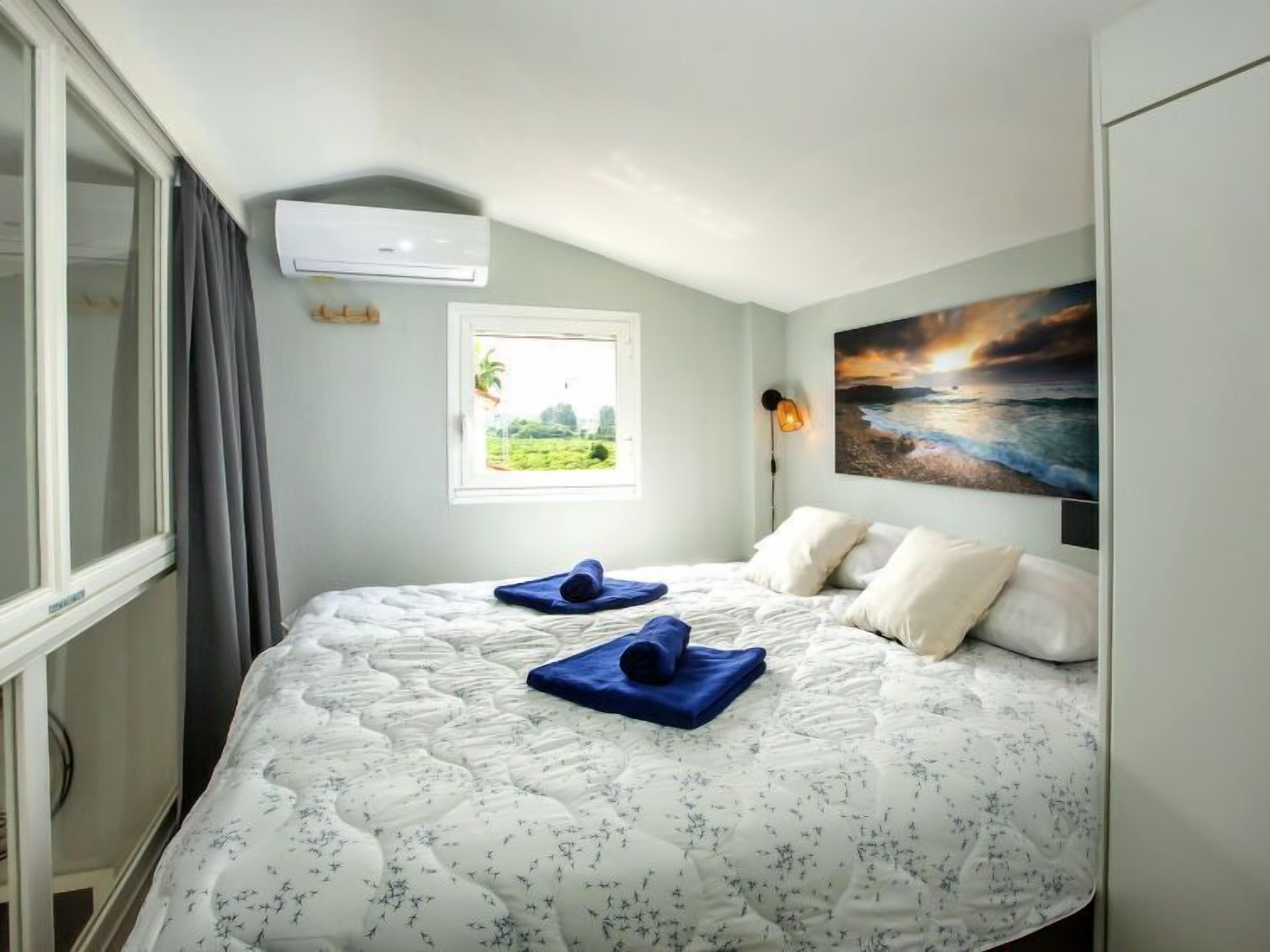 Un dormitorio con cama, una ventana con vistas a la vegetación, un equipo de aire acondicionado y un cuadro de un atardecer.
