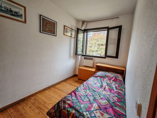 Un dormitorio pequeño con suelo de madera, cama con colcha estampada y colorida, ventana con vistas a árboles y dos cuadros en la pared.