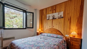 A bedroom with a wooden headboard, a bed with floral bedding, a window with a view of greenery, and two bedside lamps.