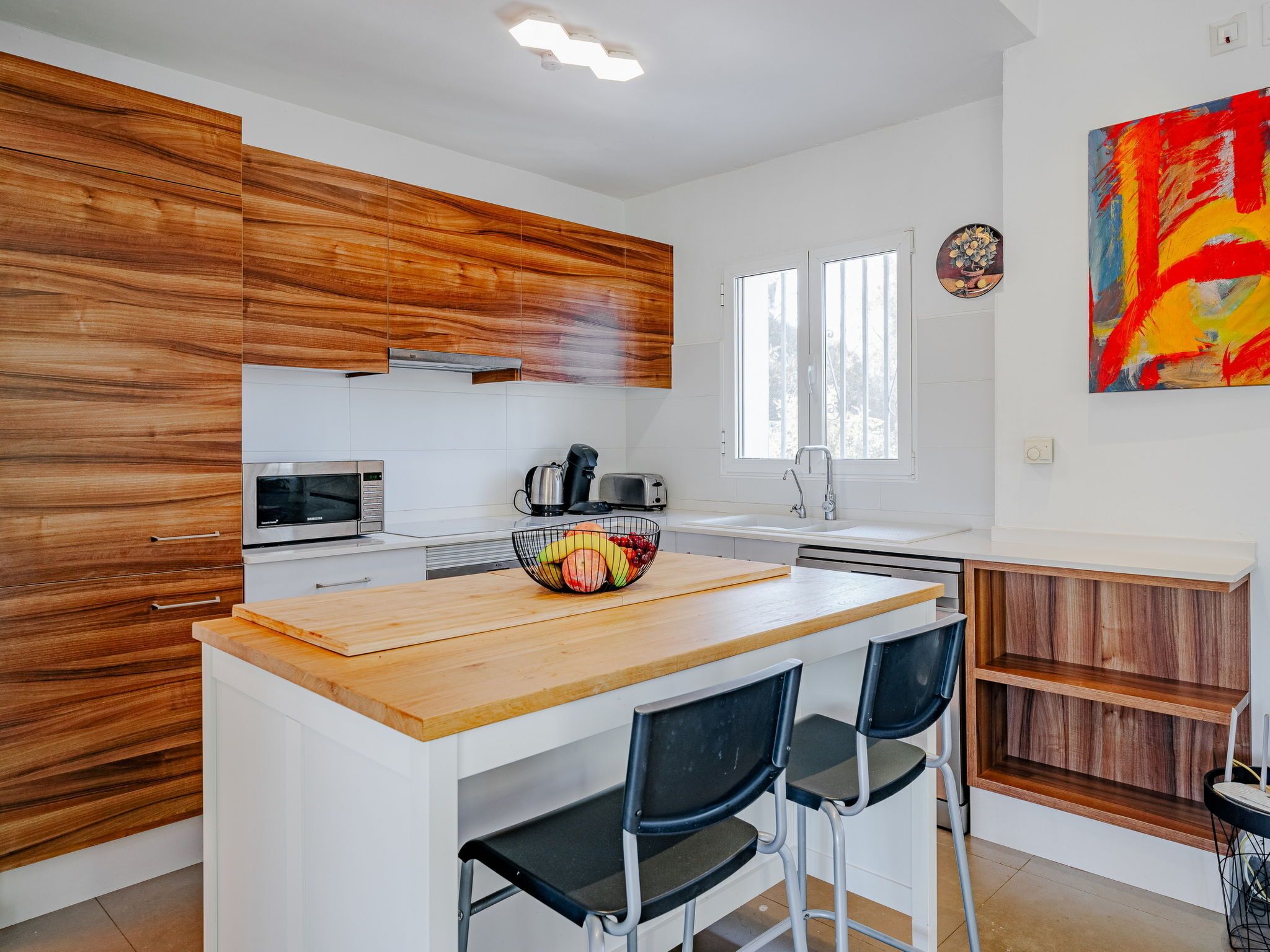 A modern kitchen with wooden cabinets, a central island with chairs, a microwave, a kettle, and a sink.