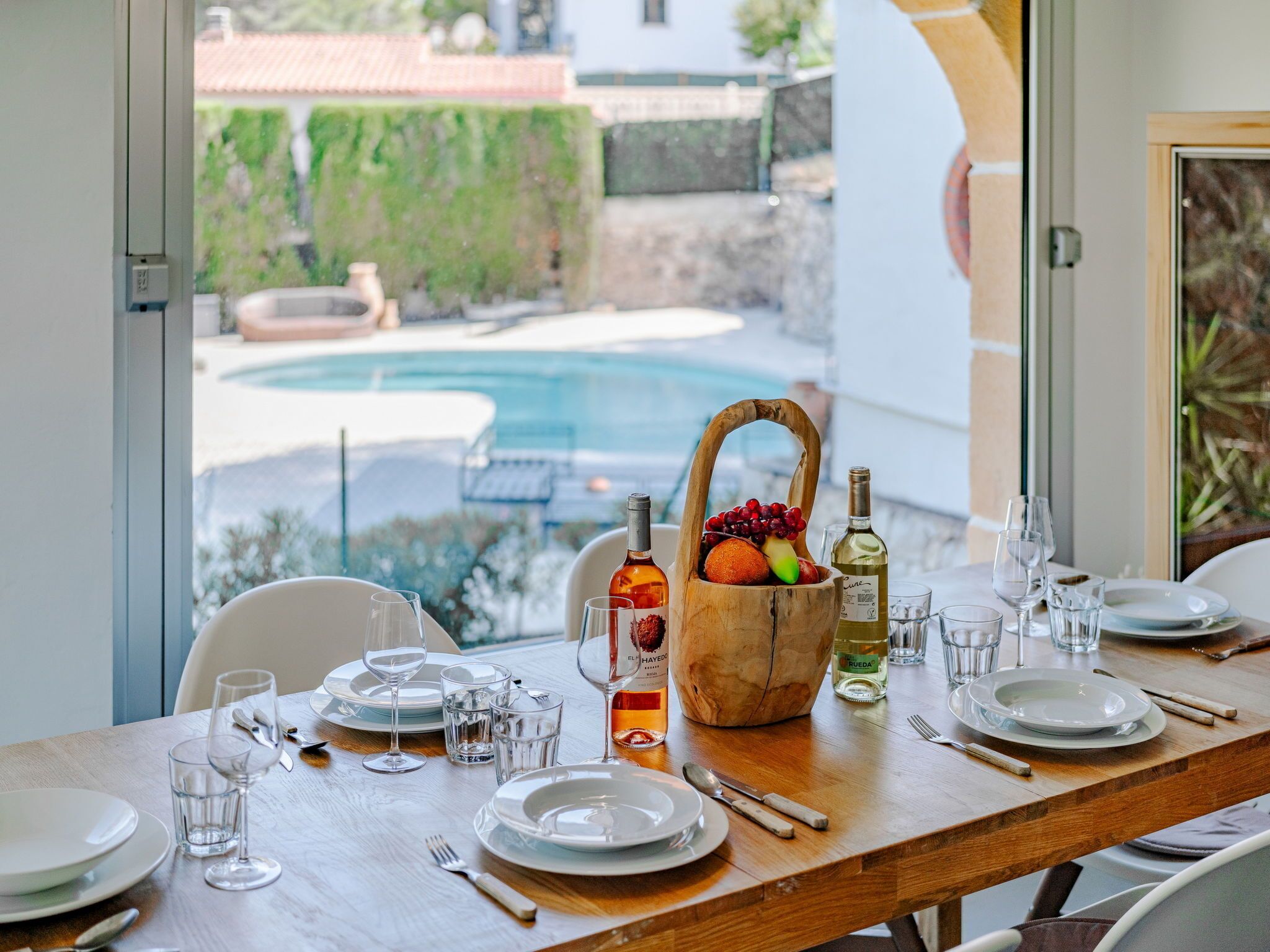A dining table set for six with plates, glasses, and cutlery, a fruit basket, and bottles of wine.