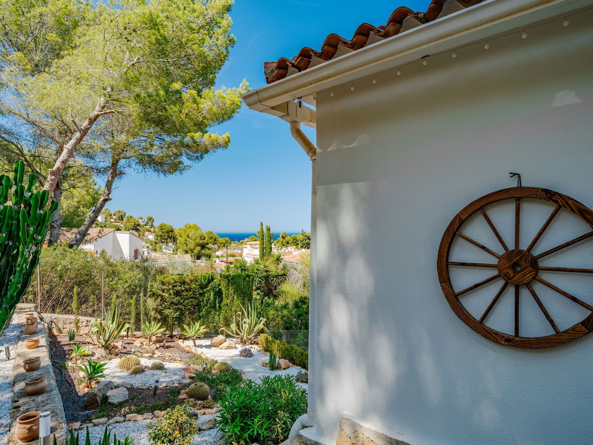A white building with a wooden wheel decoration, surrounded by a garden with various plants and a view of the sea.