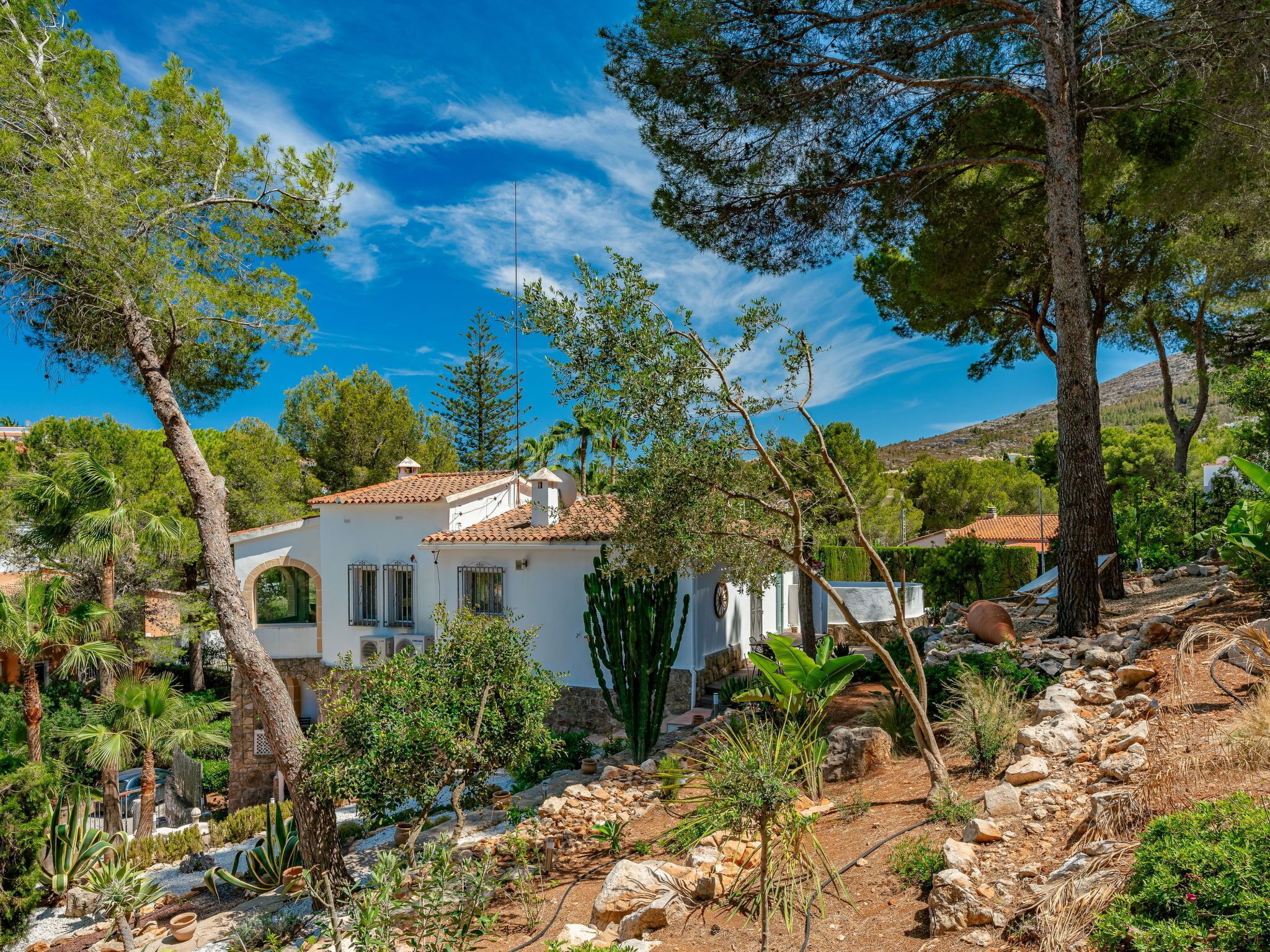 Una villa blanca con tejado de baldosas, rodeada de árboles y terreno rocoso.