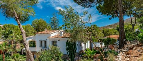 Una villa blanca con tejado de baldosas, rodeada de árboles y terreno rocoso.