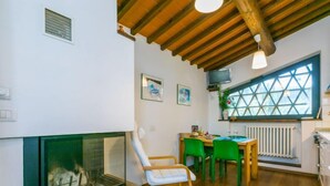 Un salon chaleureux avec une cheminée, des poutres apparentes en bois au plafond, un coin repas avec des chaises vertes et une fenêtre ornée d’un motif géométrique.