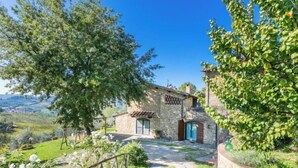 Une maison en pierre avec une allée pavée menant à un portail en bois, entourée d’une végétation luxuriante et sous un ciel bleu azur.