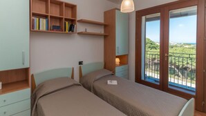 A bedroom with two beds, wooden shelving, a desk, and a balcony with a view.