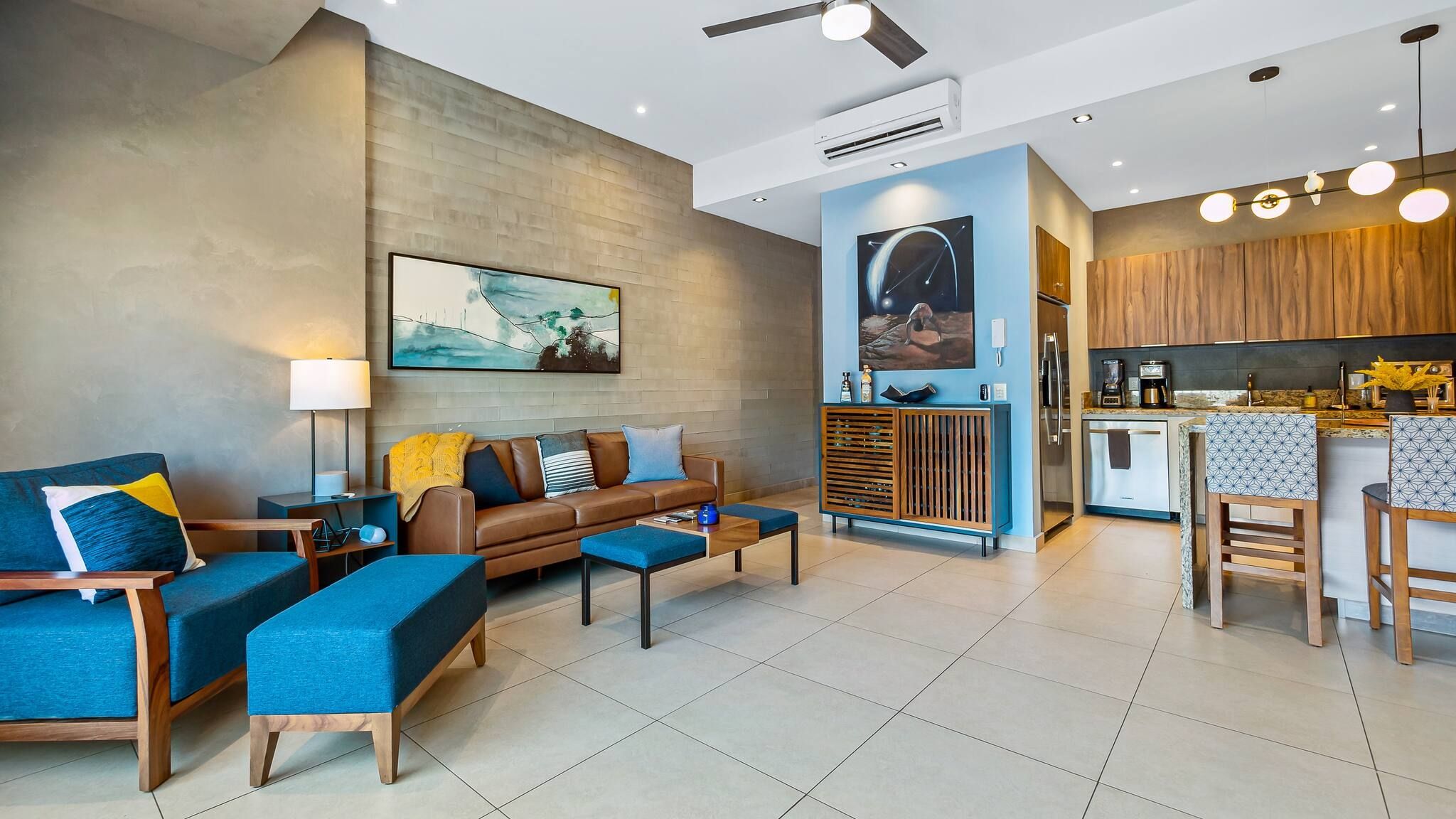 A modern living room with a sofa, coffee table, and chairs, a kitchen with wooden cabinets, and a dining area with bar stools.