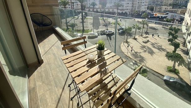 Ein Balkon mit einer hölzernen Bank und einem Blumentopf, mit Blick auf eine Stadtstraße mit Palmen und Gebäuden.