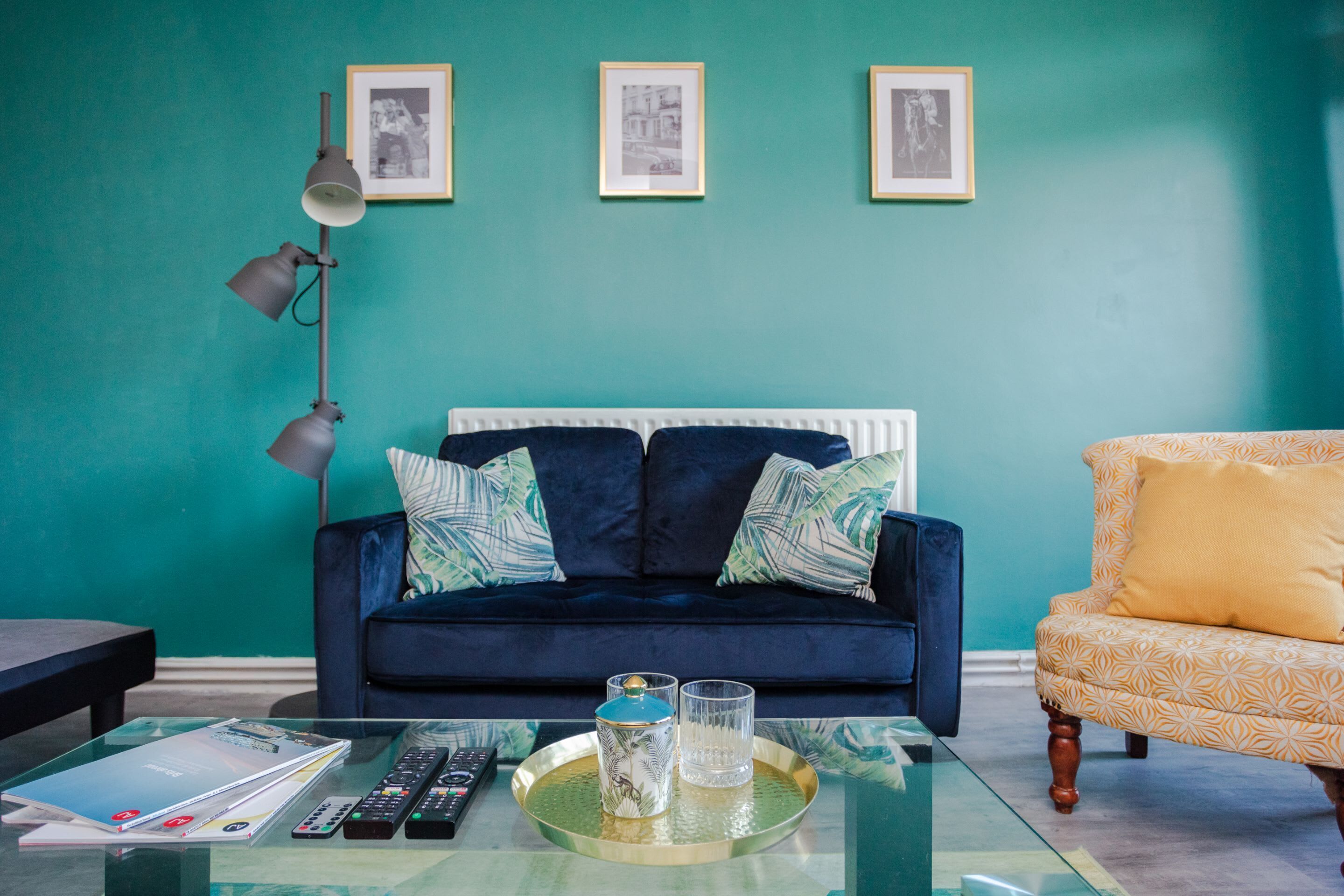A living room with a teal wall, a dark blue sofa with patterned cushions, a yellow armchair, and a glass coffee table with remote controls and a tray.