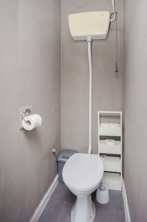A small, narrow bathroom with a toilet, a toilet paper holder, a trash bin, and a shelf with toilet paper. - Spacious 3 Bedroom Flat in the Heart of Shoreditch (London)
