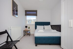 A bedroom with a bed, a bench, a radiator, a window, and a small table with a plant. - Spacious 3 Bedroom Flat in the Heart of Shoreditch (London)