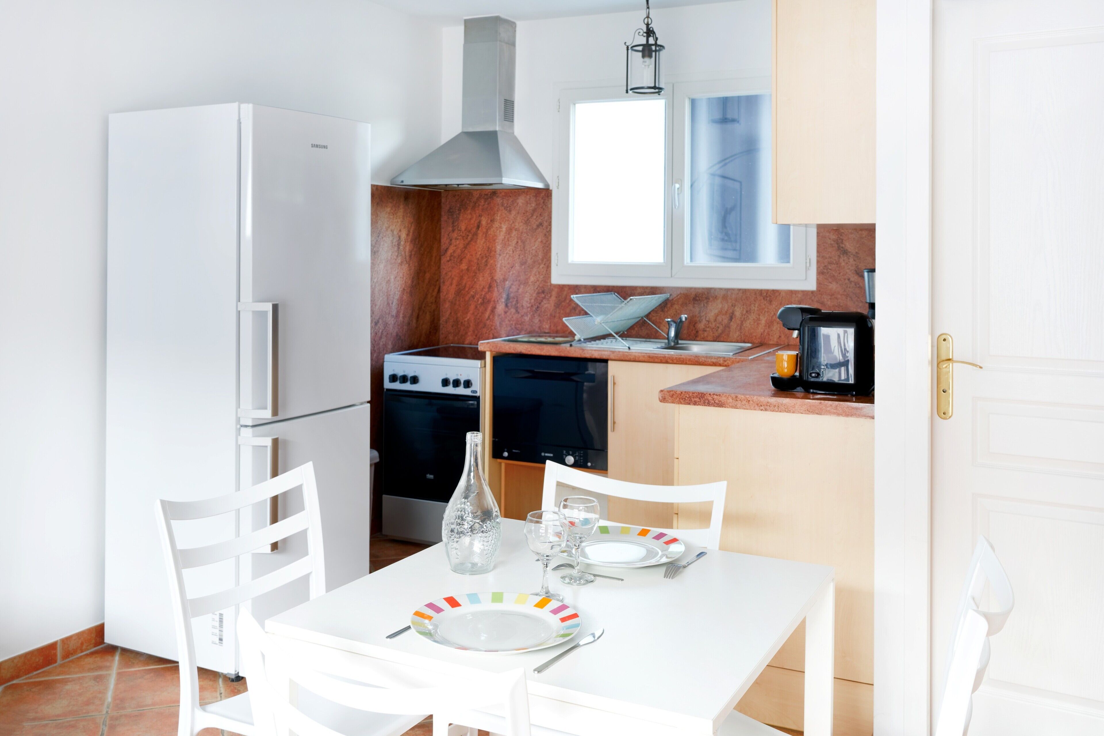 Een kleine keuken met witte apparaten, een eettafel gedekt voor twee personen, en een raam boven de gootsteen.