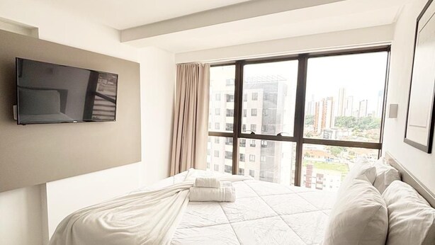 A modern hotel room with a large bed, a flat-screen TV mounted on the wall, and a city view through the large window. - Manaira Apart Hotel (João Pessoa)