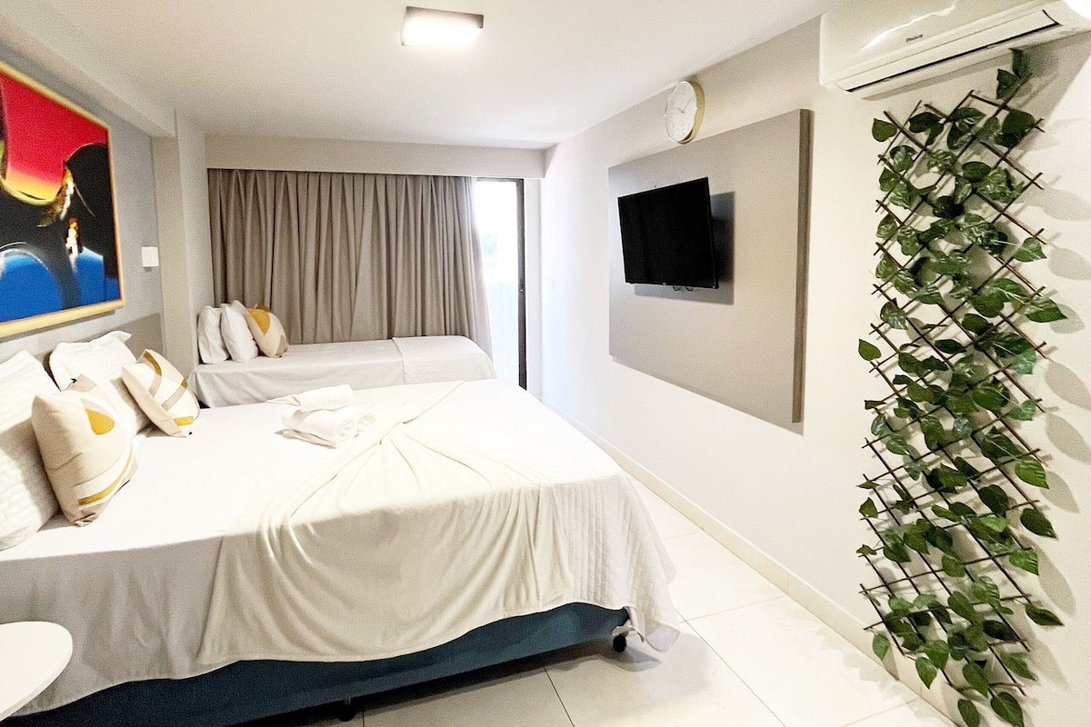 A hotel room with a bed, a flat-screen TV, an air conditioning unit, and a wall-mounted plant.