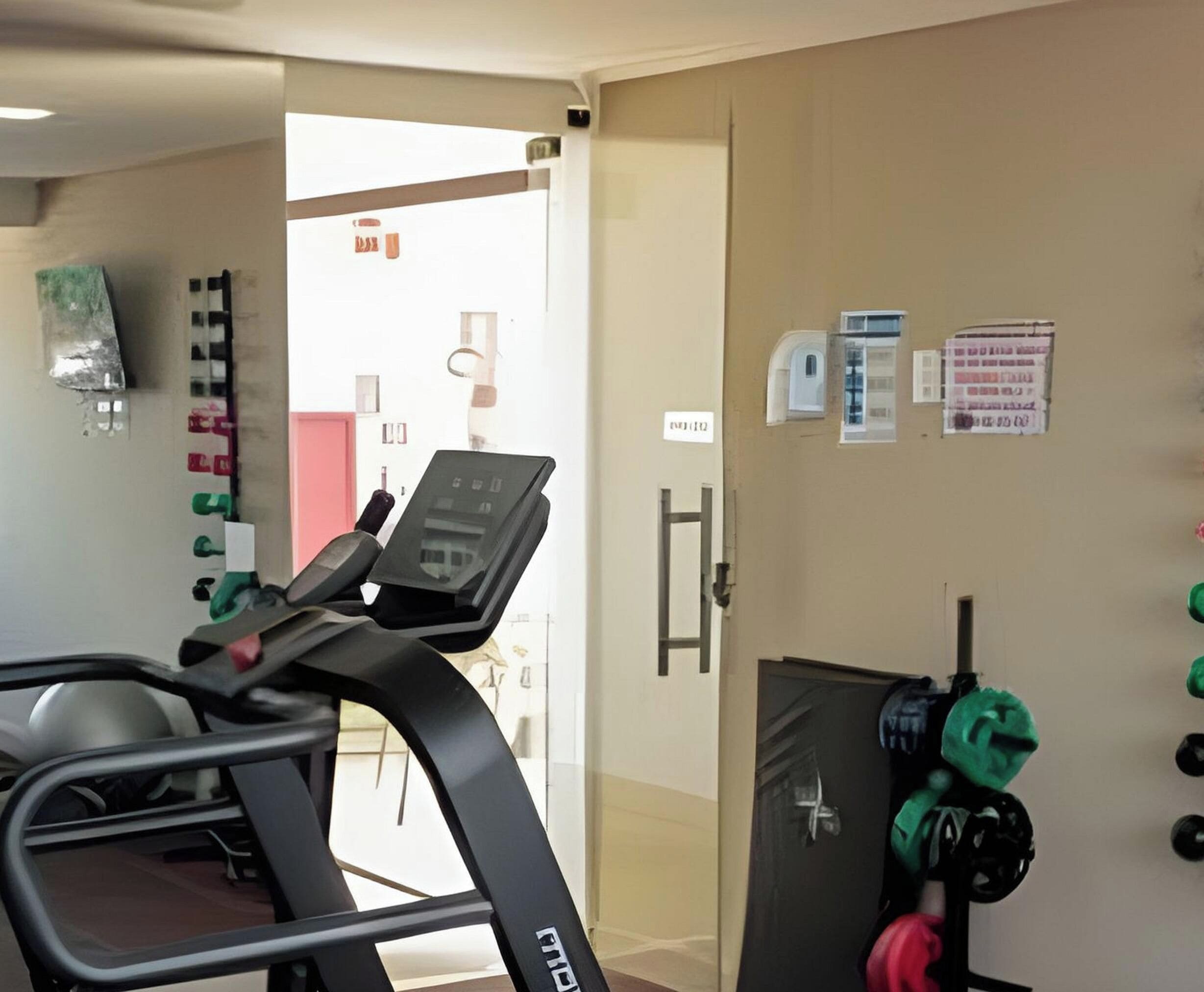 Une salle de sport équipée de matériel d’exercice, d’un tapis de course et d’un mur où sont affichés divers panneaux et avis.