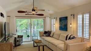 A spacious living room with a large sofa, a TV, and a ceiling fan. There is a sliding glass door leading to a balcony with a view of trees.