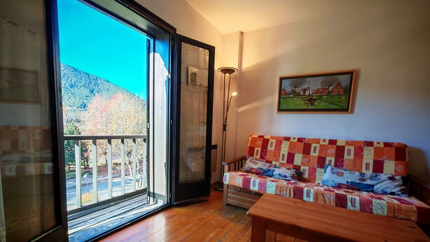 Ein Zimmer mit Holzboden, einem Sofa mit farbenfroher Stegdecke, einer Bank, einem Bild an der Wand und einer Glastür, die auf einen Balkon mit Blick auf Bäume und ein Gebäude führt.