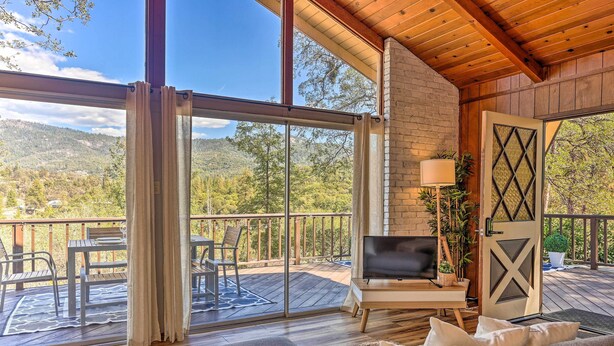 A spacious living area with a large window, a sofa, a TV, and a wooden deck with outdoor furniture. - Lovely Oakhurst Cabin w/ Deck & Mtn Views! (Oakhurst)