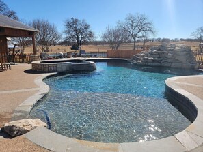 A clear swimming pool with a stone edge, surrounded by a wooden deck and a stone wall. - Private Retreat with Pond, Pool and Hot Tub (Boyd)