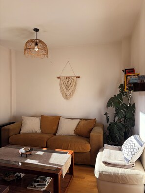 A living room with a brown sofa, a coffee table, a hanging macrame wall piece, a plant, and a bookshelf with books.
