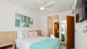 Een moderne hotelkamer met een groot bed, een plafondventilator, een schilderij aan de muur en een houten deur die naar een andere kamer leidt.