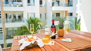 Een houten tafel gedekt met champagne, een ananas, glazen en een mand met brood op een balkon met uitzicht op een modern gebouw.