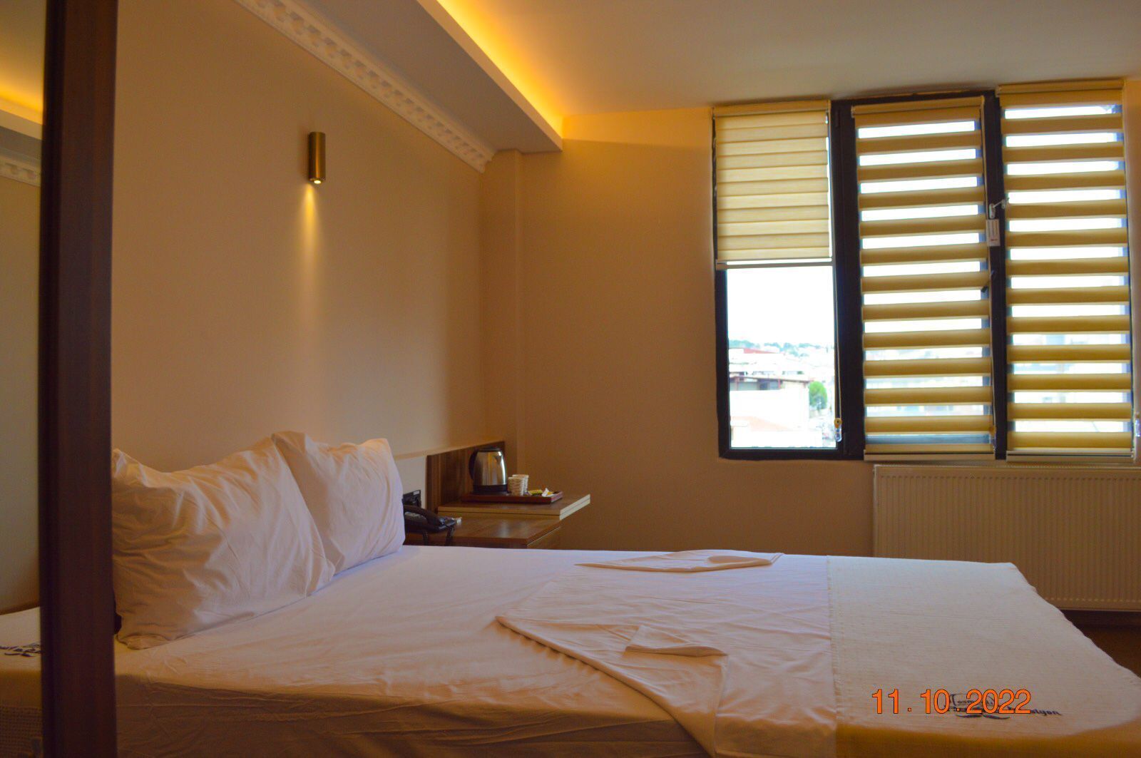 A hotel room with a bed, bedside table, lamp, and window with blinds.