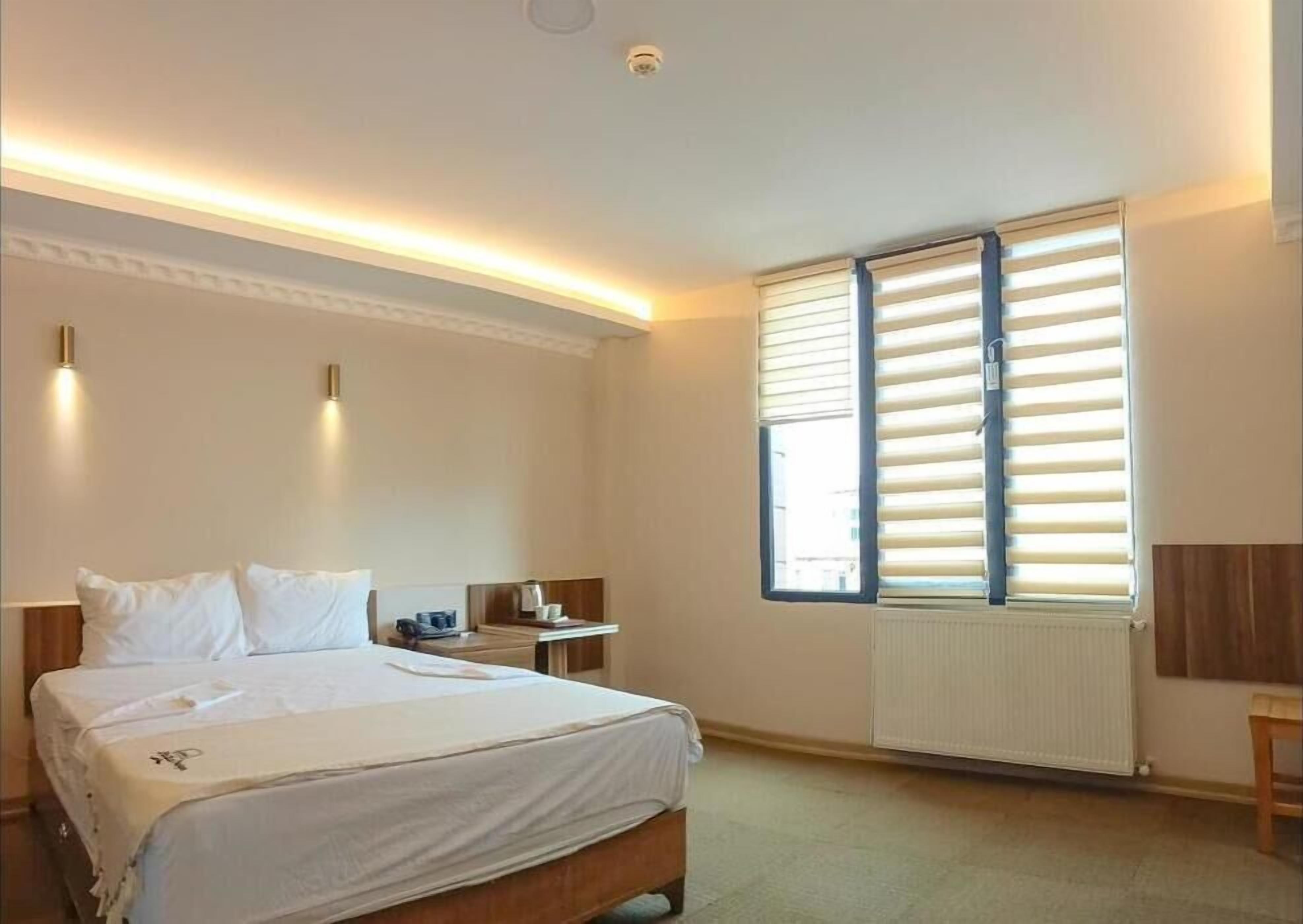 A hotel room with a bed, bedside tables, a window with blinds, and a radiator.