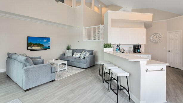 A modern living room with a grey sofa, a coffee table, and a TV mounted on the wall. There is a staircase leading to an upper level.