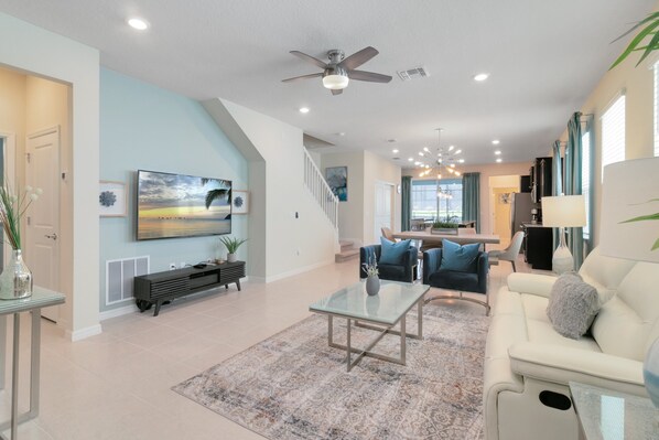 A spacious living room with a large flat-screen TV, a ceiling fan, and a staircase leading to the upper floor.