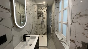 Ein modernes Badezimmer mit einem weißen Waschbecken, einem Spiegel mit Beleuchtung, einer Dusche und einem Fenster.