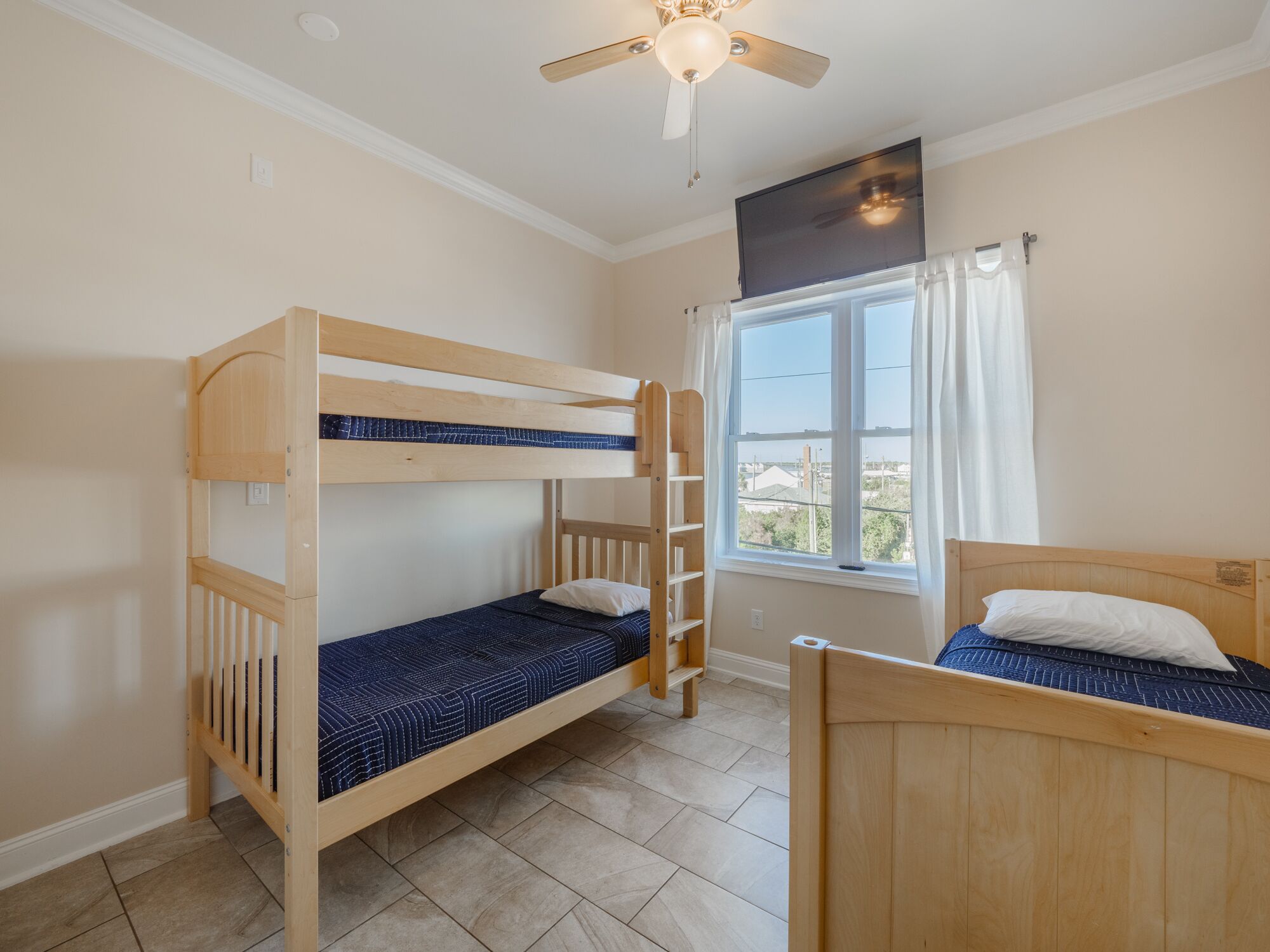 Une chambre avec deux lits superposés, un ventilateur de plafond, une fenêtre avec des rideaux et un téléviseur fixé au mur.