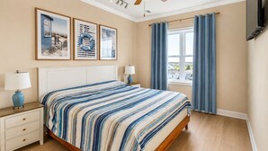 A bedroom with a bed, nightstands, a ceiling fan, a window with curtains, and wall-mounted artwork. - Big Blue -Mini-Resort, Movie Theater, Pools, Hot Tub, Beach Front, Sleeps 28! (Surf City)